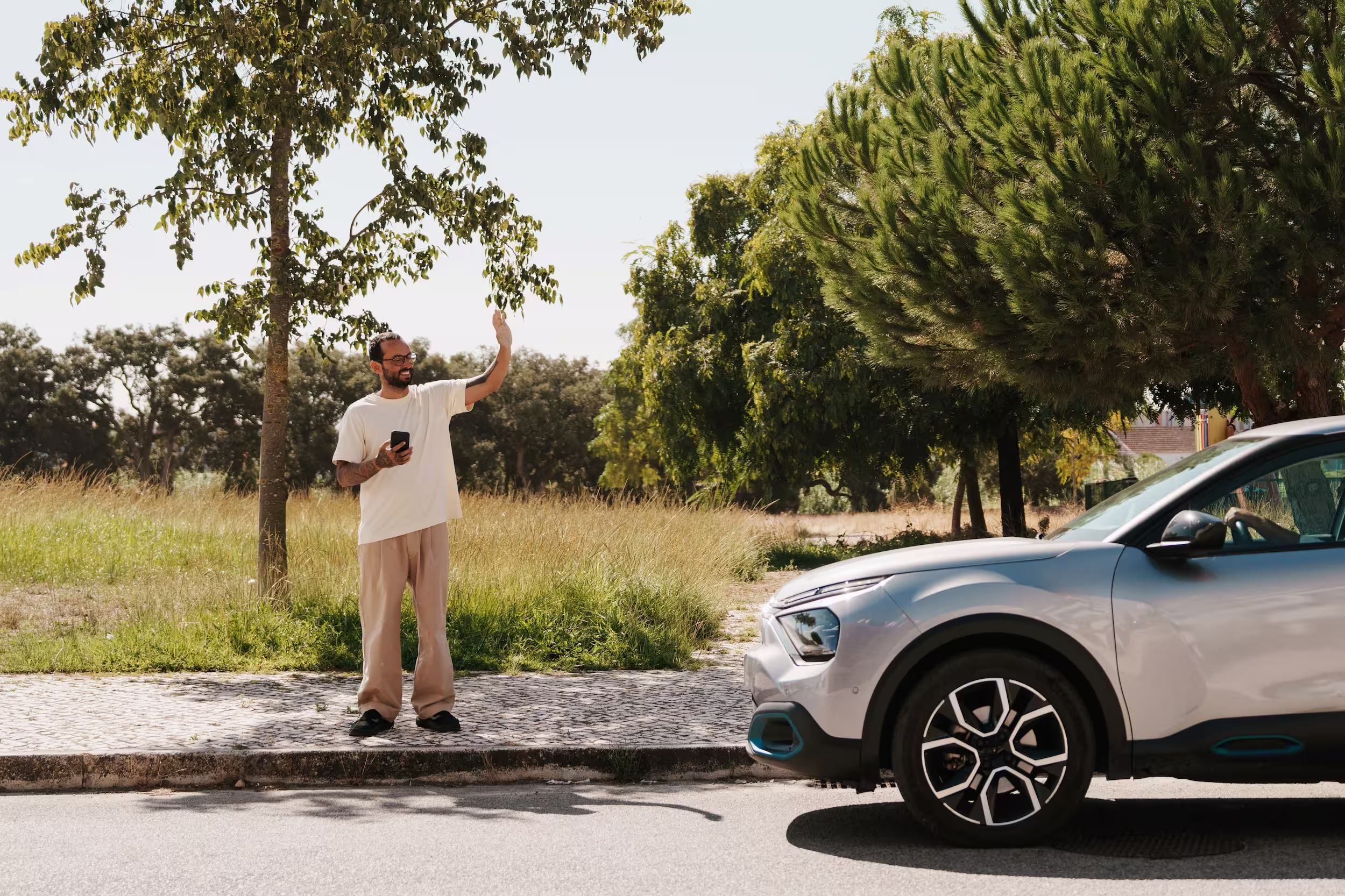A man wearing glasses stands on a sidewalk and waves at a gray car on a sunny day in a green environment.