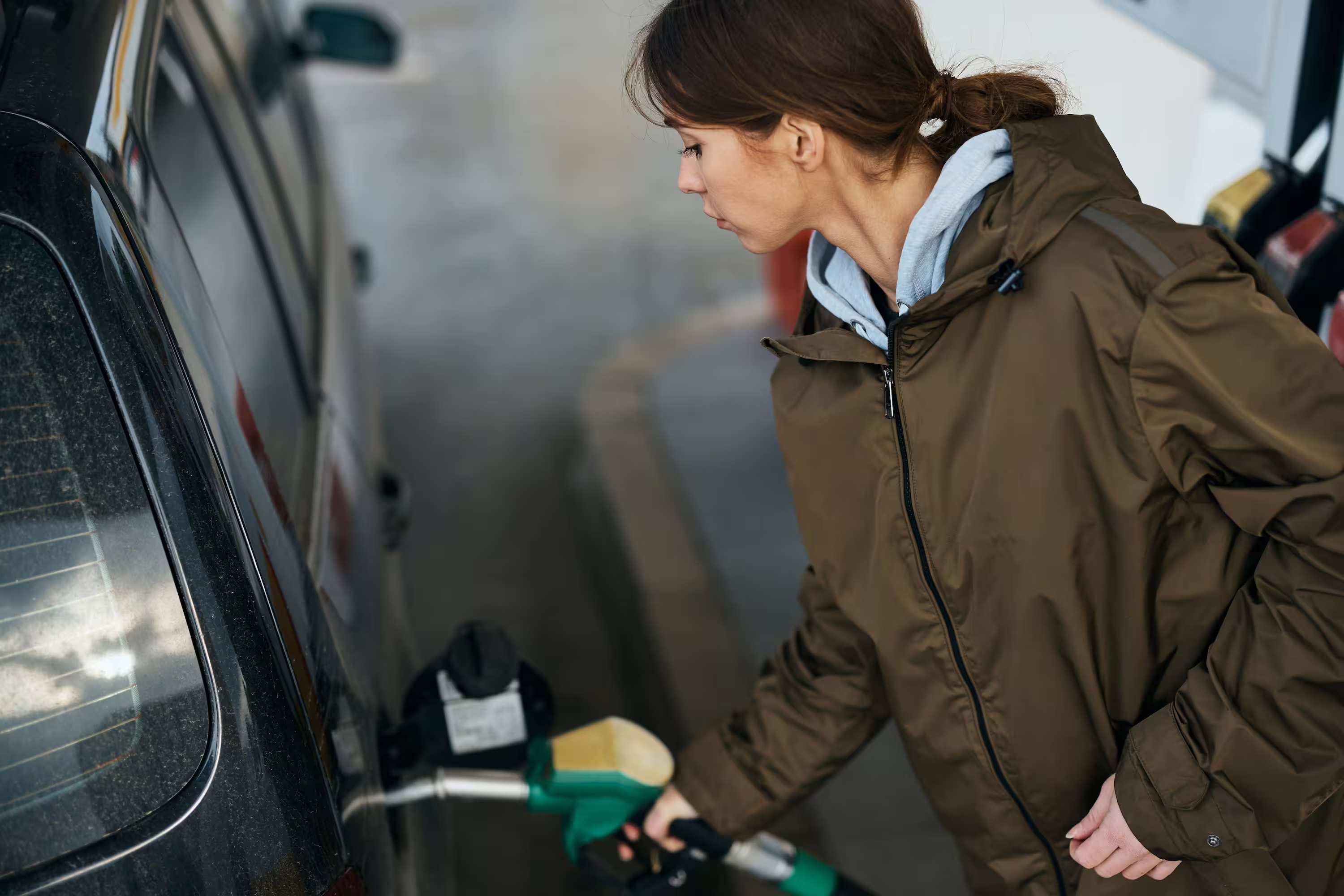 Woman in a brown jacket refueling a black car at a gas station.